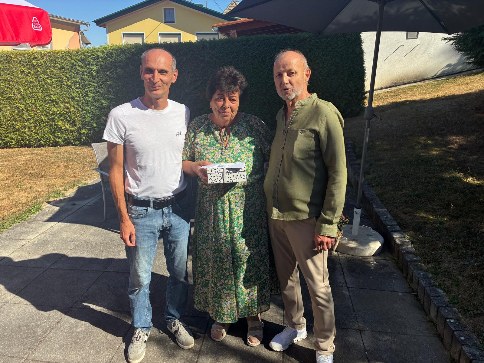 Drei Personen stehen im Garten, eine Frau hält eine Geschenkbox.