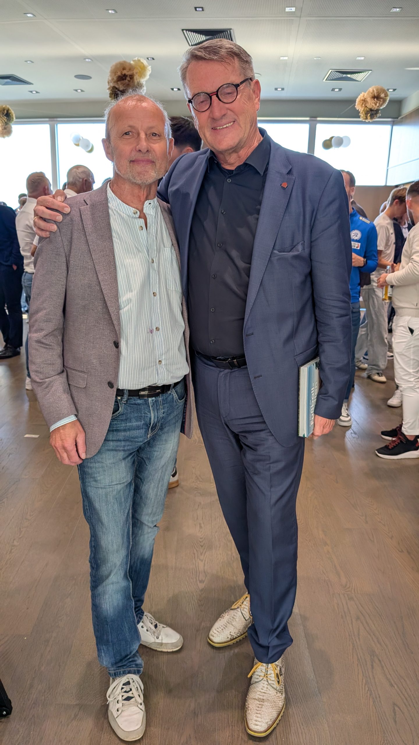 Zwei Männer in einem Innenraum, einer mit blauem Anzug, der andere mit Sakko und Jeans